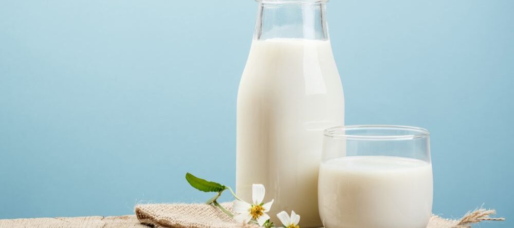 A glass and bottle of milk sit on a hessian patch, on a wooden table beside delicate flowers