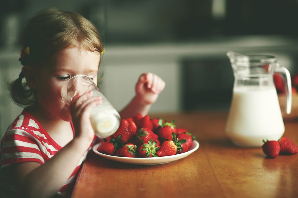 A child drinks from a glass of milk with a plate of vibrant strawberries in front of her.