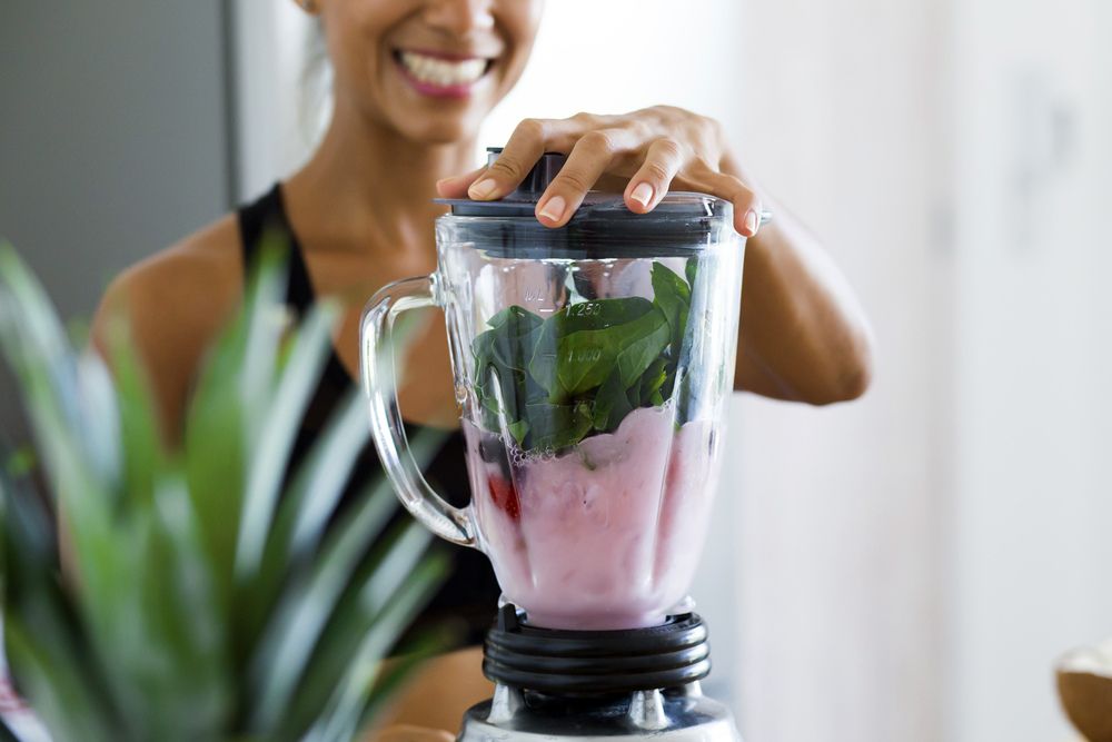 A smiling woman adds greens, milk and berries to a smoothie maker.